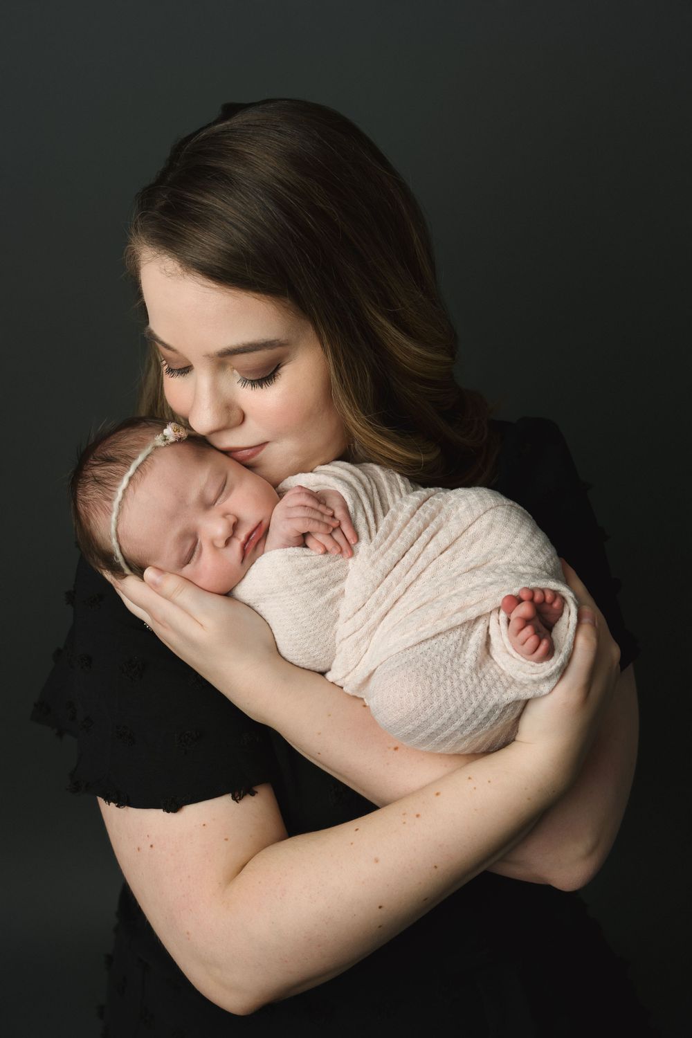 Loving mother in black dress kissing sleeping swaddled newborn baby girl against dark background.