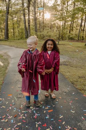 Two young children in burgundy graduation gowns sharing a joyful moment on a path surrounded by trees.