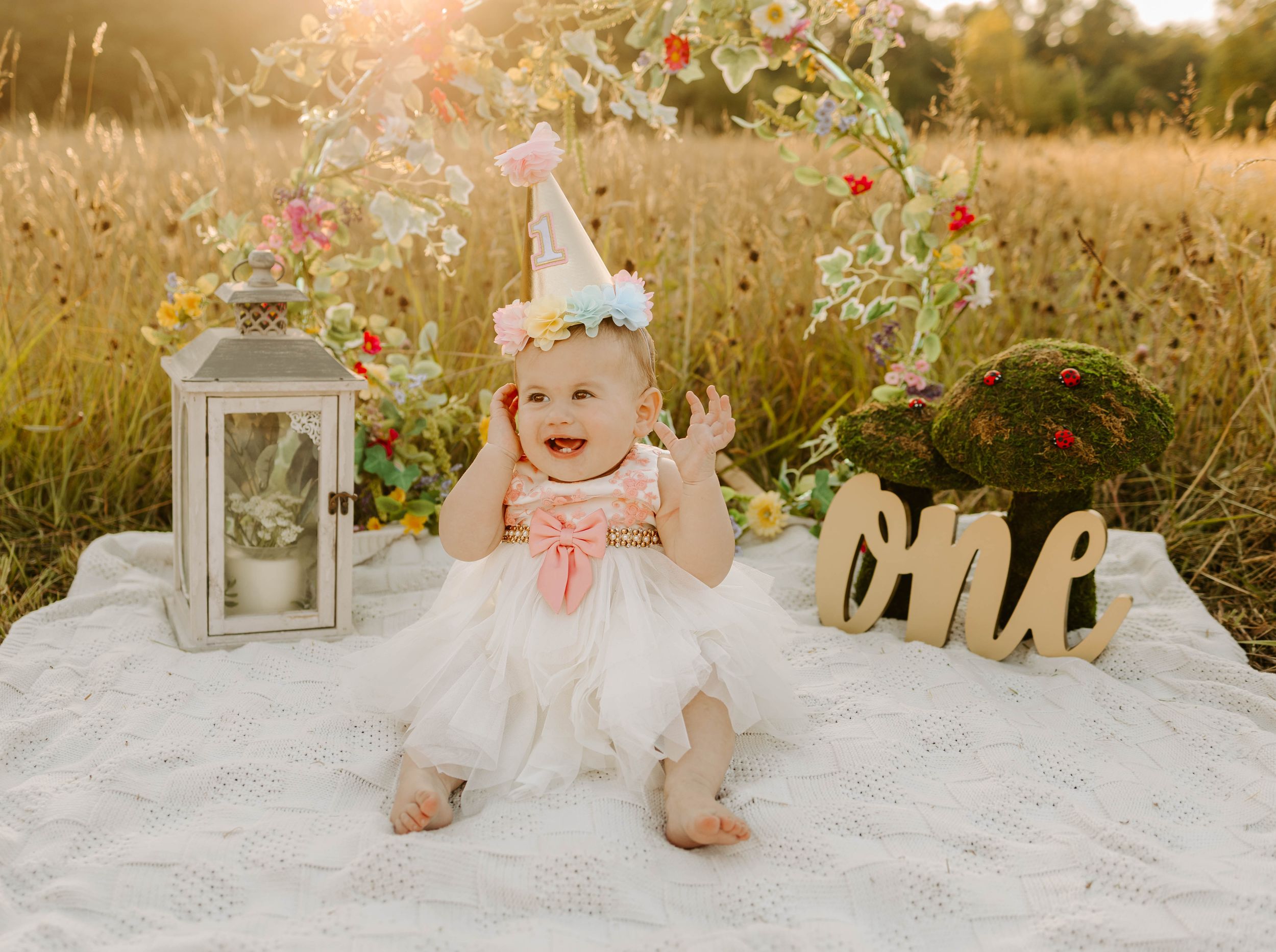 Baby in white tulle dress sits on blanket with lantern and wooden letters at sunset.