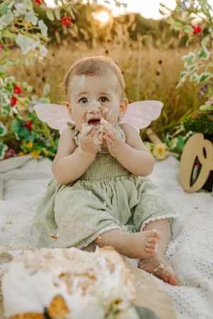 Whimsical cake smash photography with fairy wings and floral decorations in sunset field.