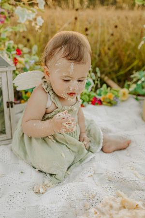 Messy cake smash photo with sage green dress and white lace blanket outdoors.