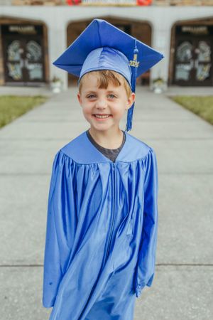 Young graduate wearing bright blue cap and gown smiles outside school building.
