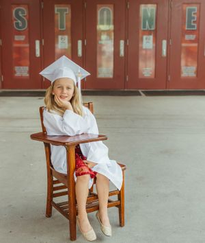 Student in white graduation attire sits in wooden chair against red lockers spelling STONE.