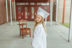 Graduate in white cap and gown poses near red lockers.