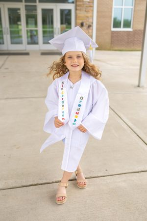 Graduate in white cap and gown with decorative sash poses cheerfully outside a school building.