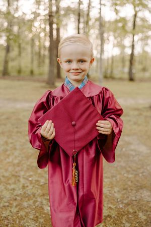 Young graduate in burgundy gown holding graduation cap in wooded outdoor setting.
