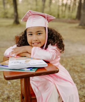 Young graduate in pink cap and gown smiling while leaning on desk with books outdoors.