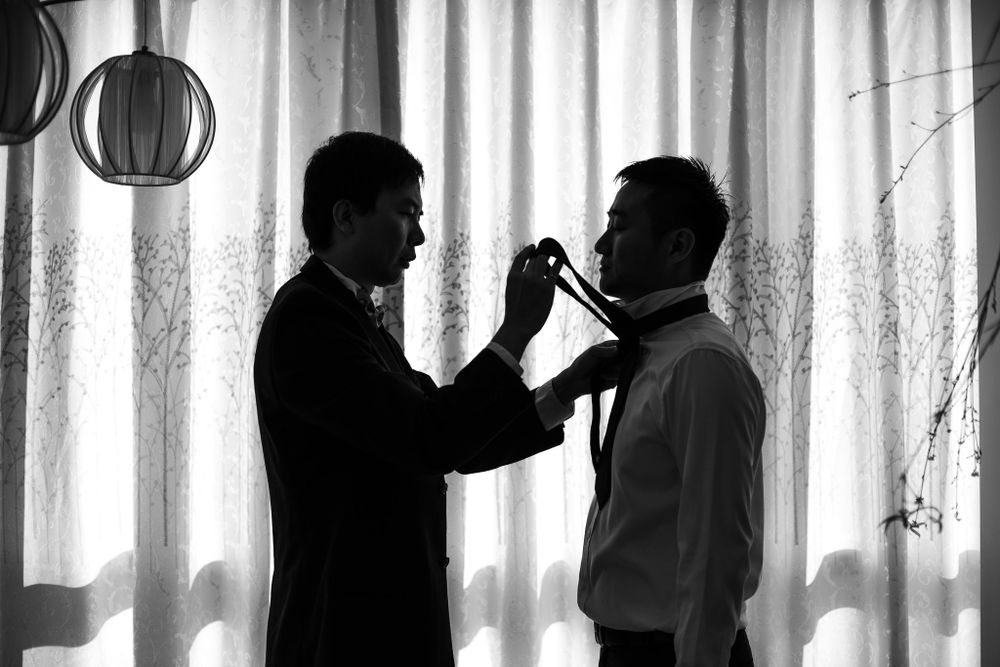 Silhouette of two figures in formal wear adjusting a tie against a white curtain backdrop.