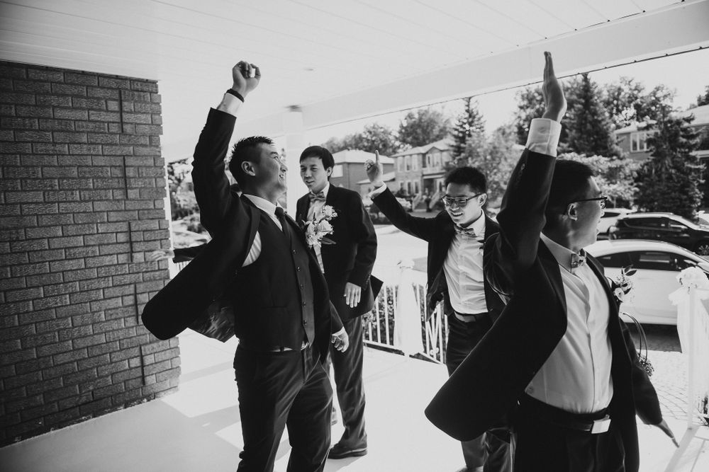 Groomsmen celebrating with raised arms outside a brick building on a sunny day.