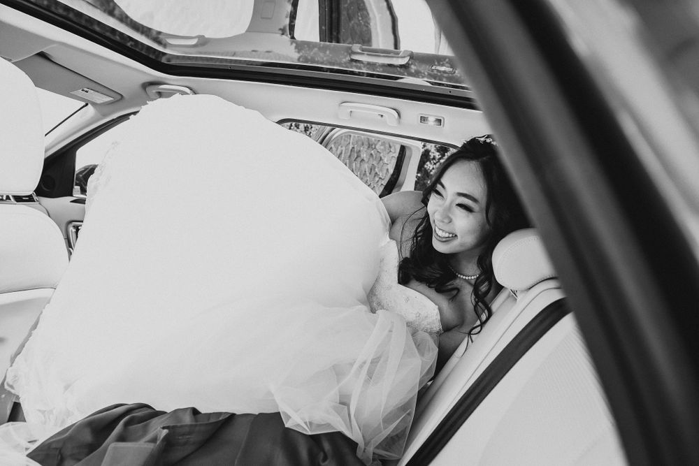 Black and white photo of a bride in white dress entering a car.