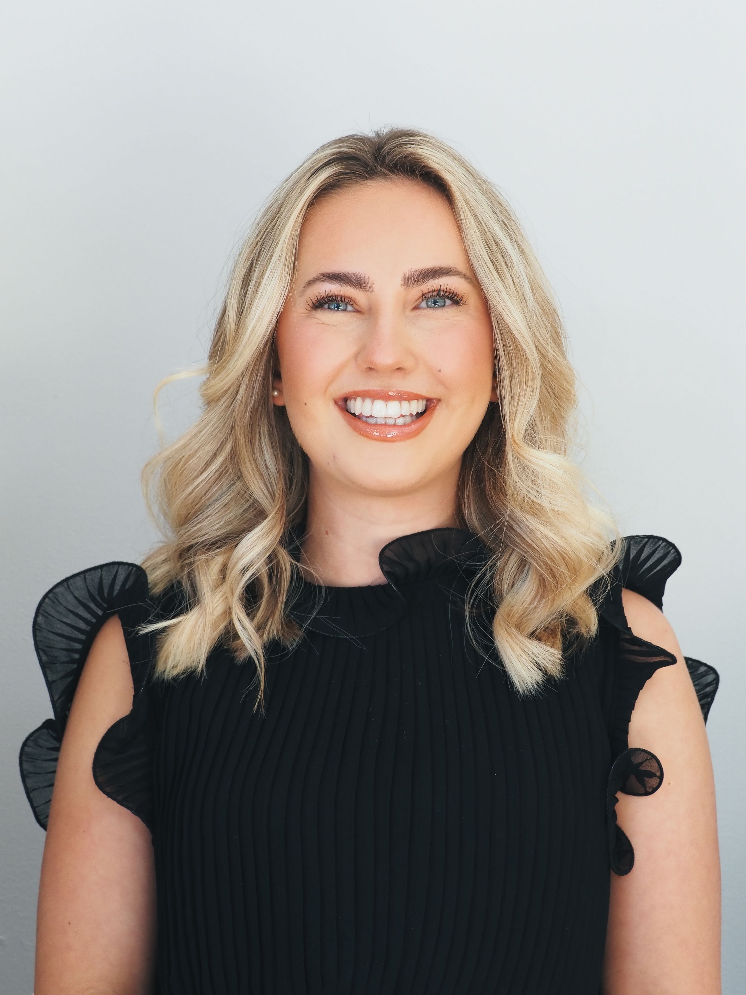 Professional headshot of someone with wavy blonde hair wearing a black top with ruffled sleeves against a light background.