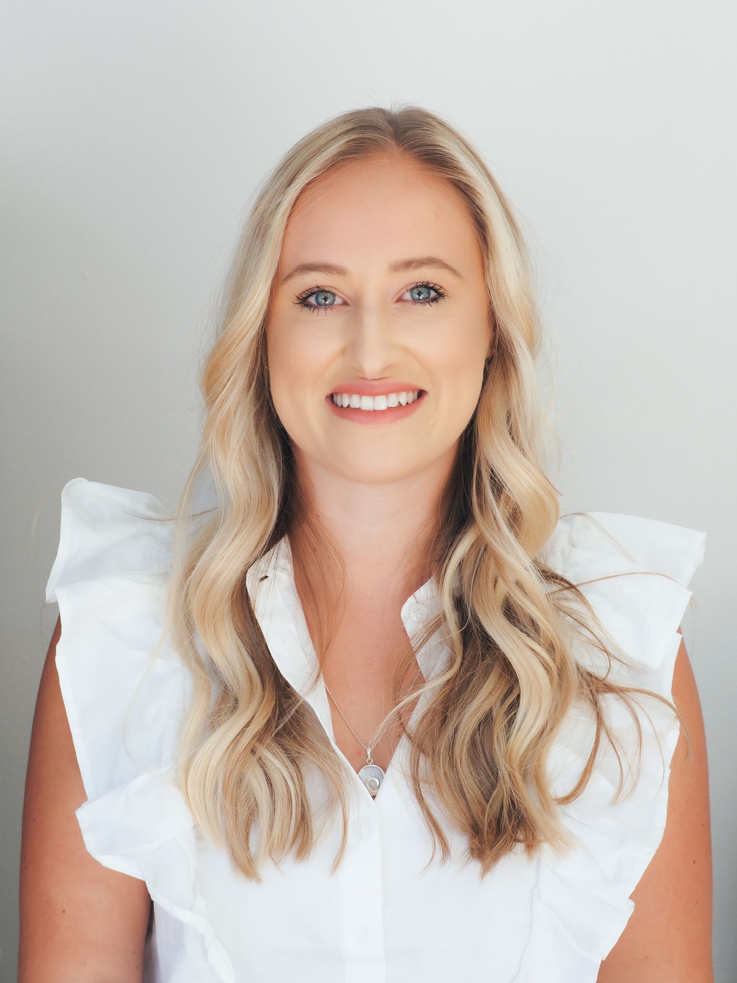 Professional headshot of someone with wavy blonde hair wearing a white ruffled sleeveless top against a light gray background.