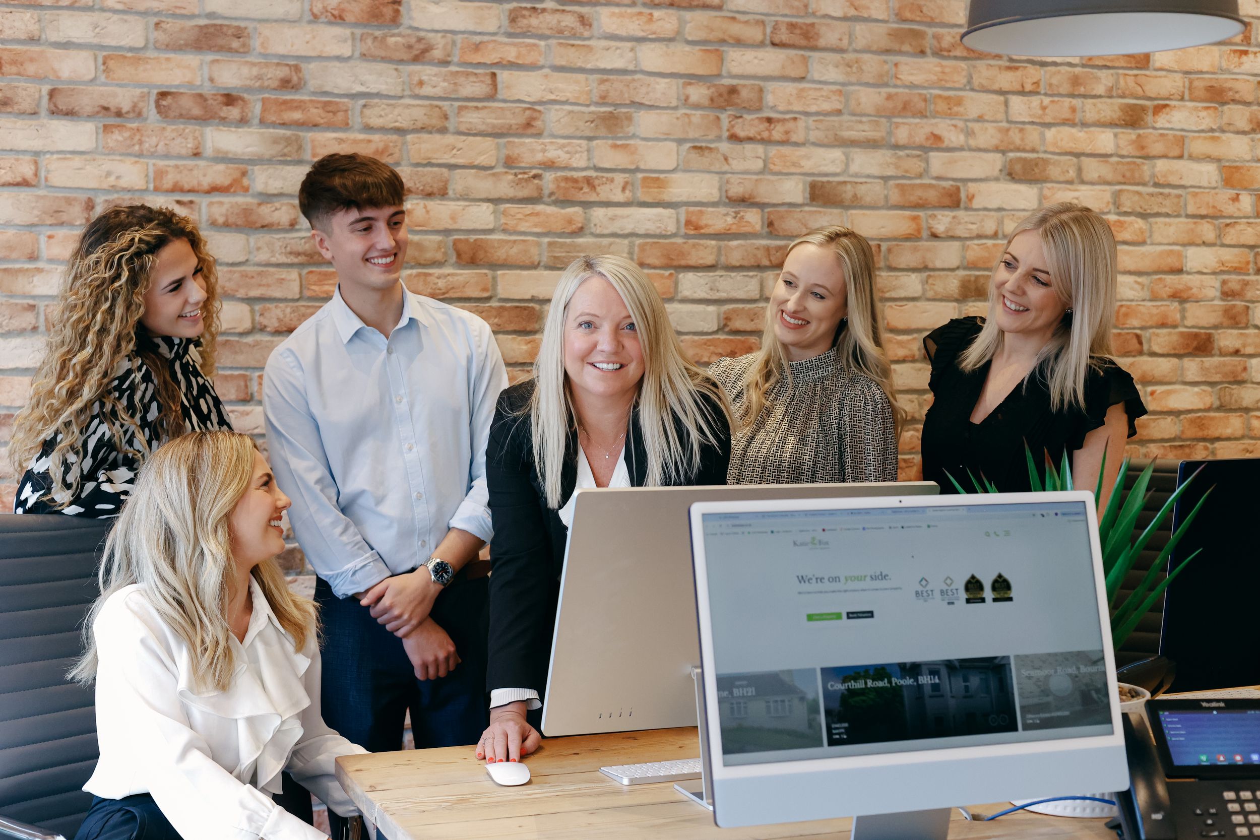 A group of people collaborating at a desk while looking at a laptop computer in a modern brick-walled office.