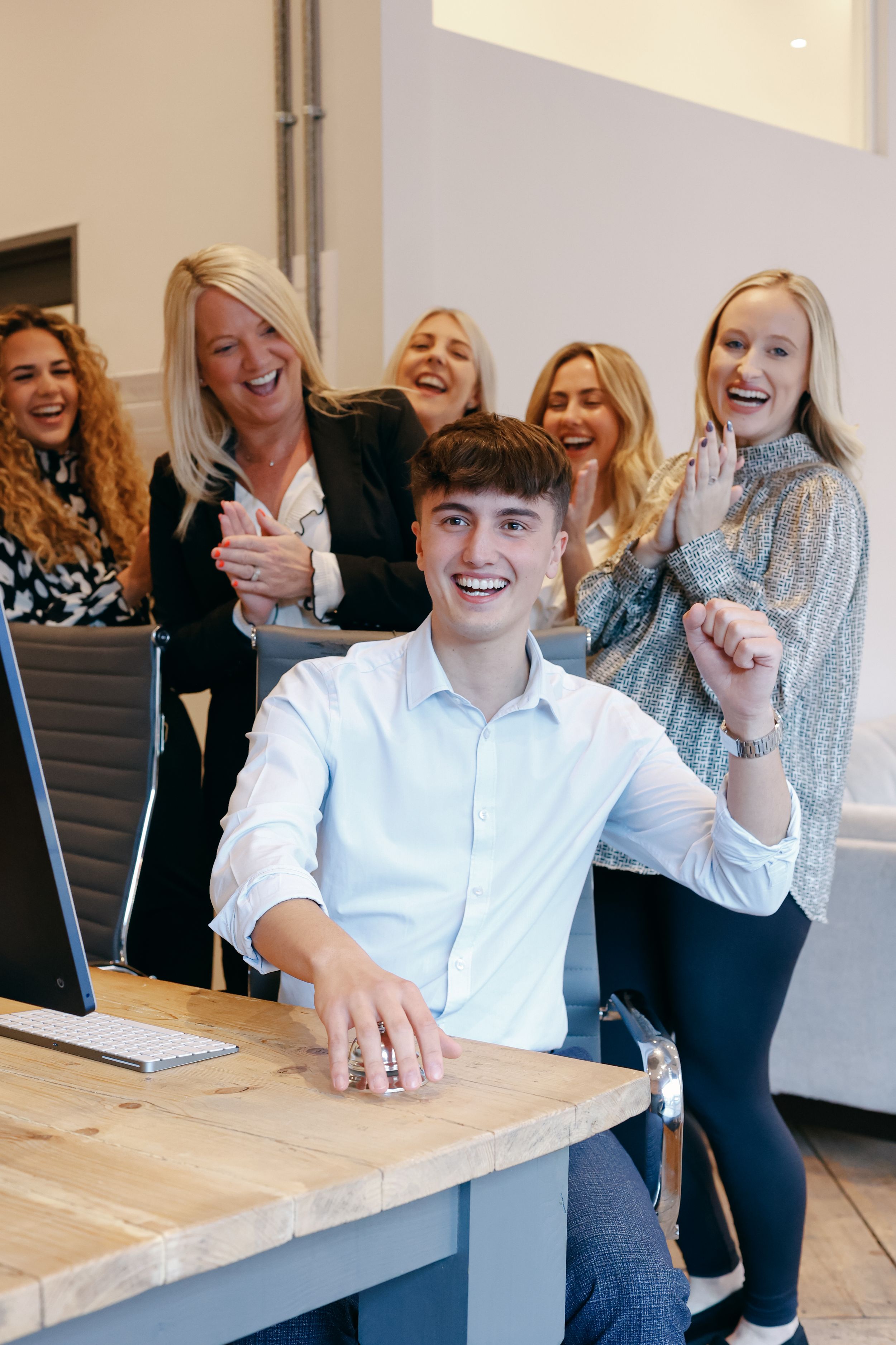 Group of colleagues celebrating and laughing together at wooden desk in modern workspace.