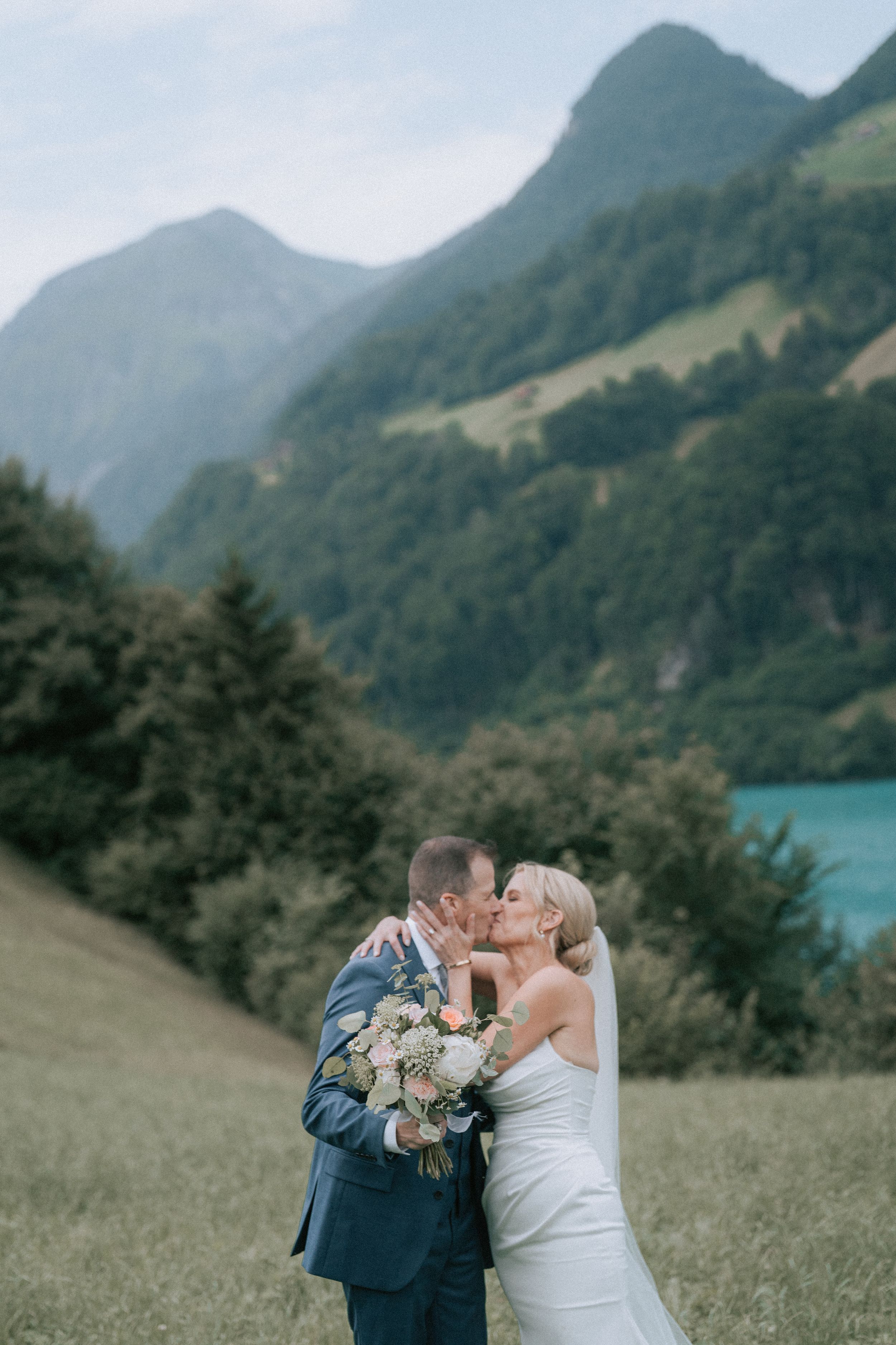 A couple shares a loving moment on a grassy hillside overlooking a turquoise alpine lake surrounded by mountains.