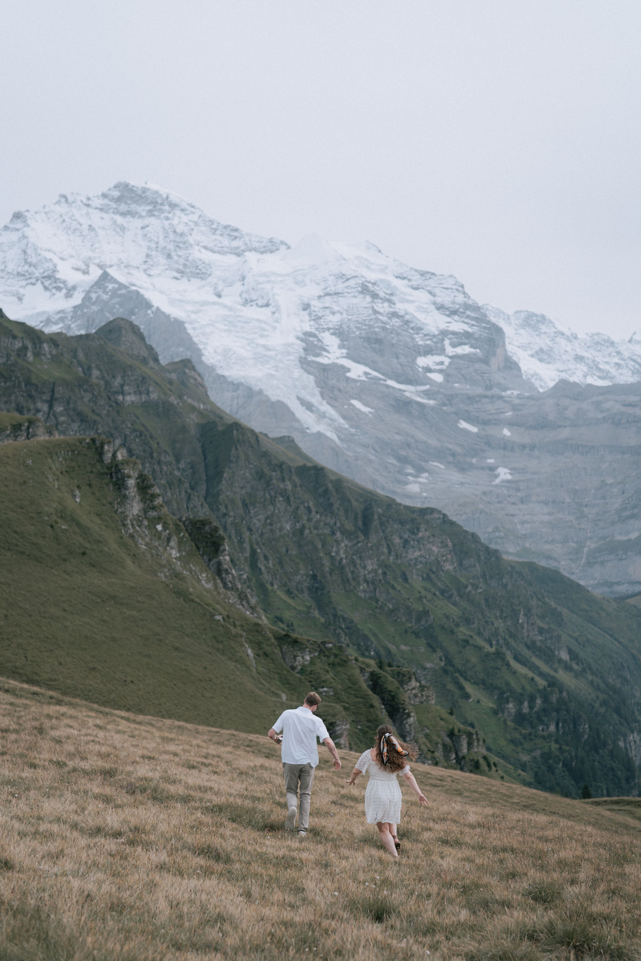 Two people walk hand in hand through an alpine meadow with snow-capped mountain peaks in the Swiss Alps background.