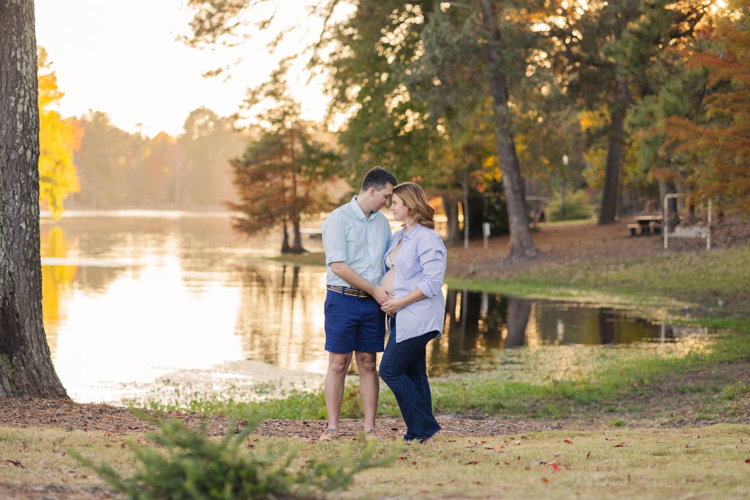 An expectant mother in a cream dress stands thoughtfully in a sunlit forest setting.