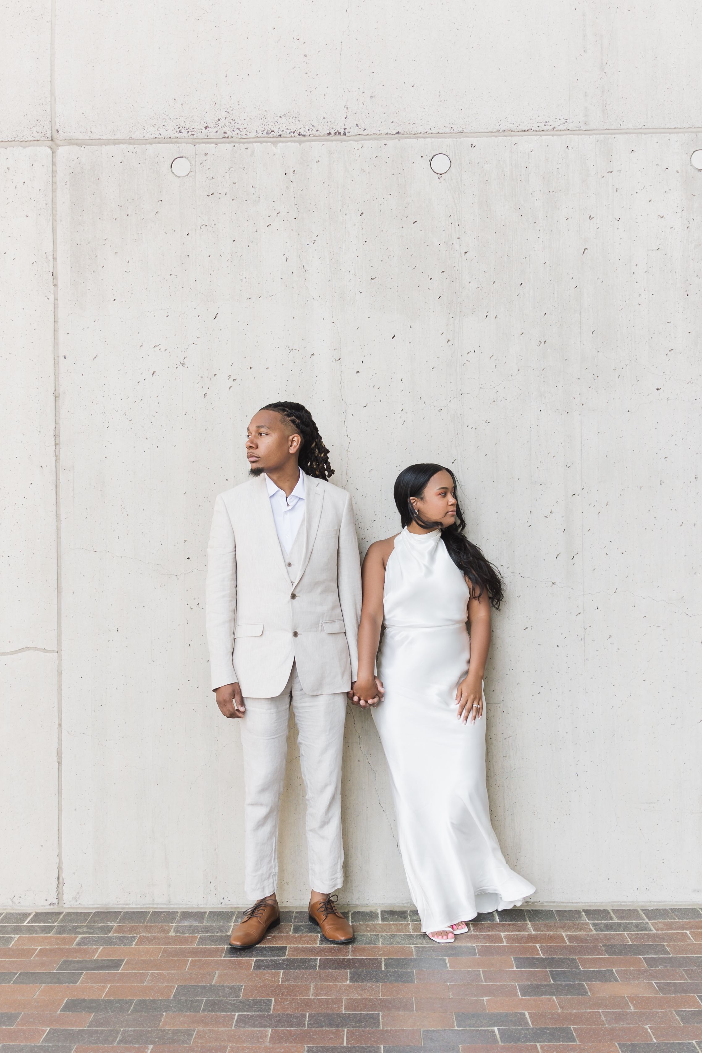 Minimalist wedding portrait of couple in white attire against concrete wall.