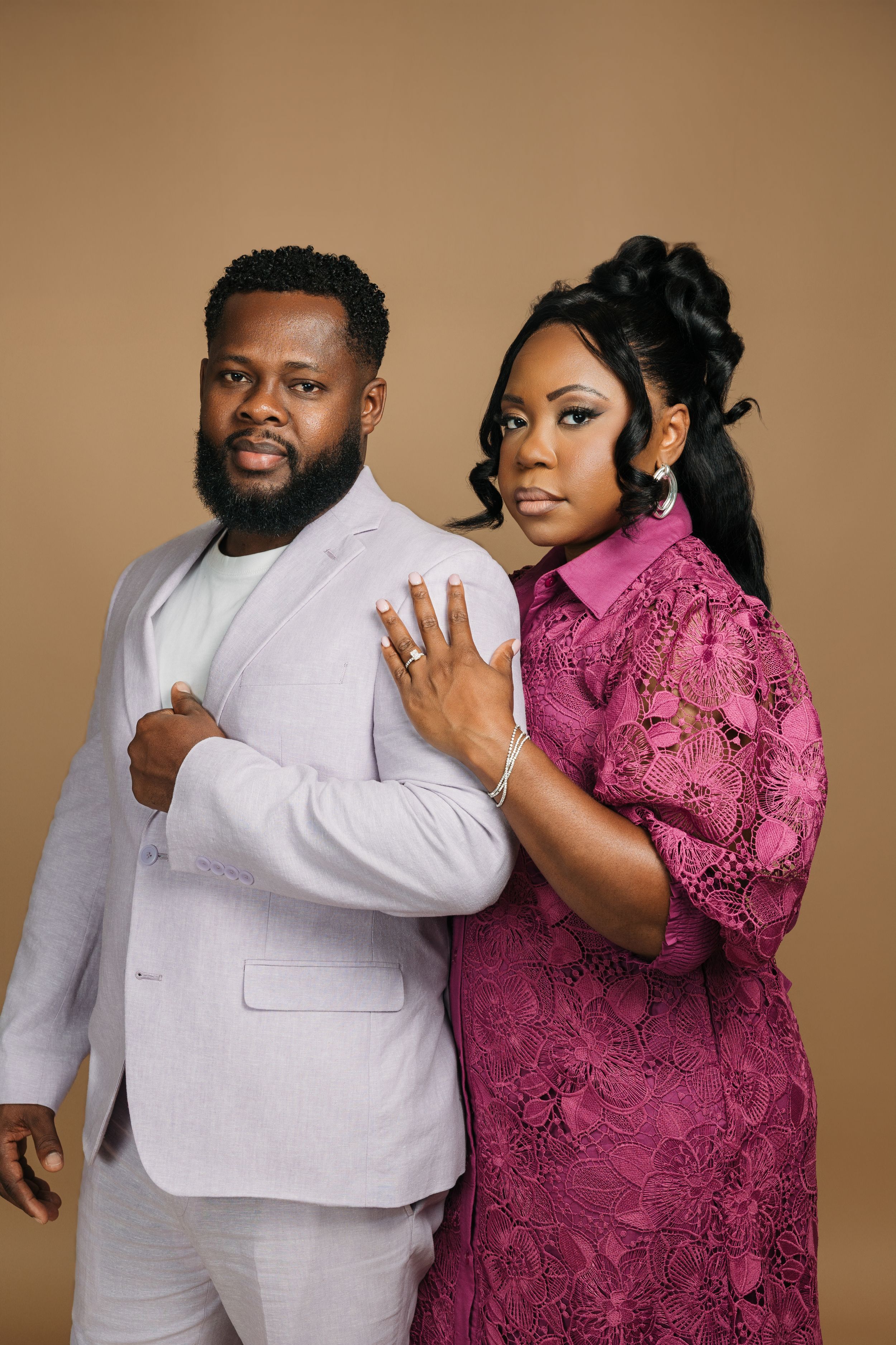 A couple in white and magenta formal wear pose playfully together in a studio against a brown backdrop.