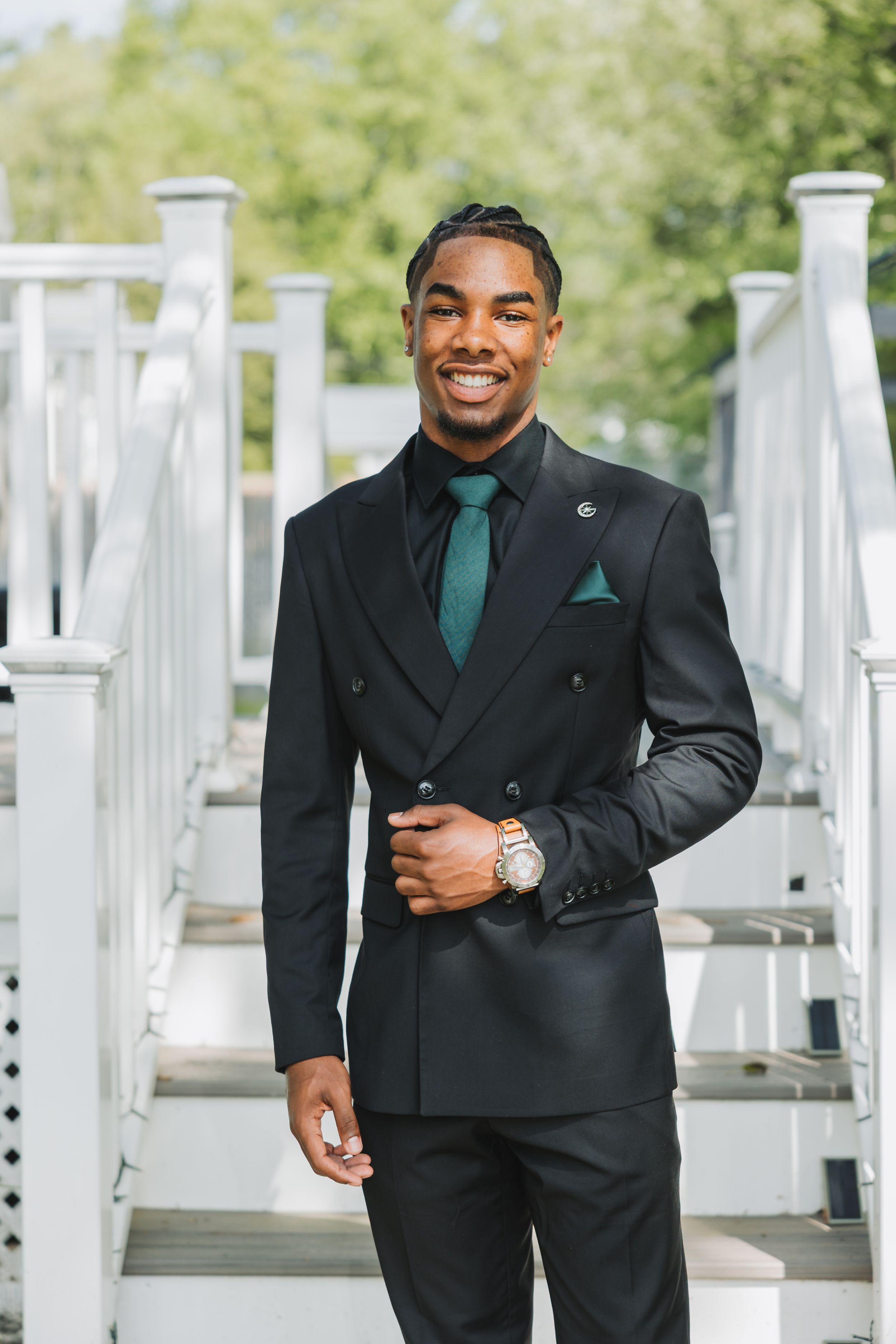 A well-dressed person in a double-breasted black suit with green tie and pocket square smiles while standing on white stairs.