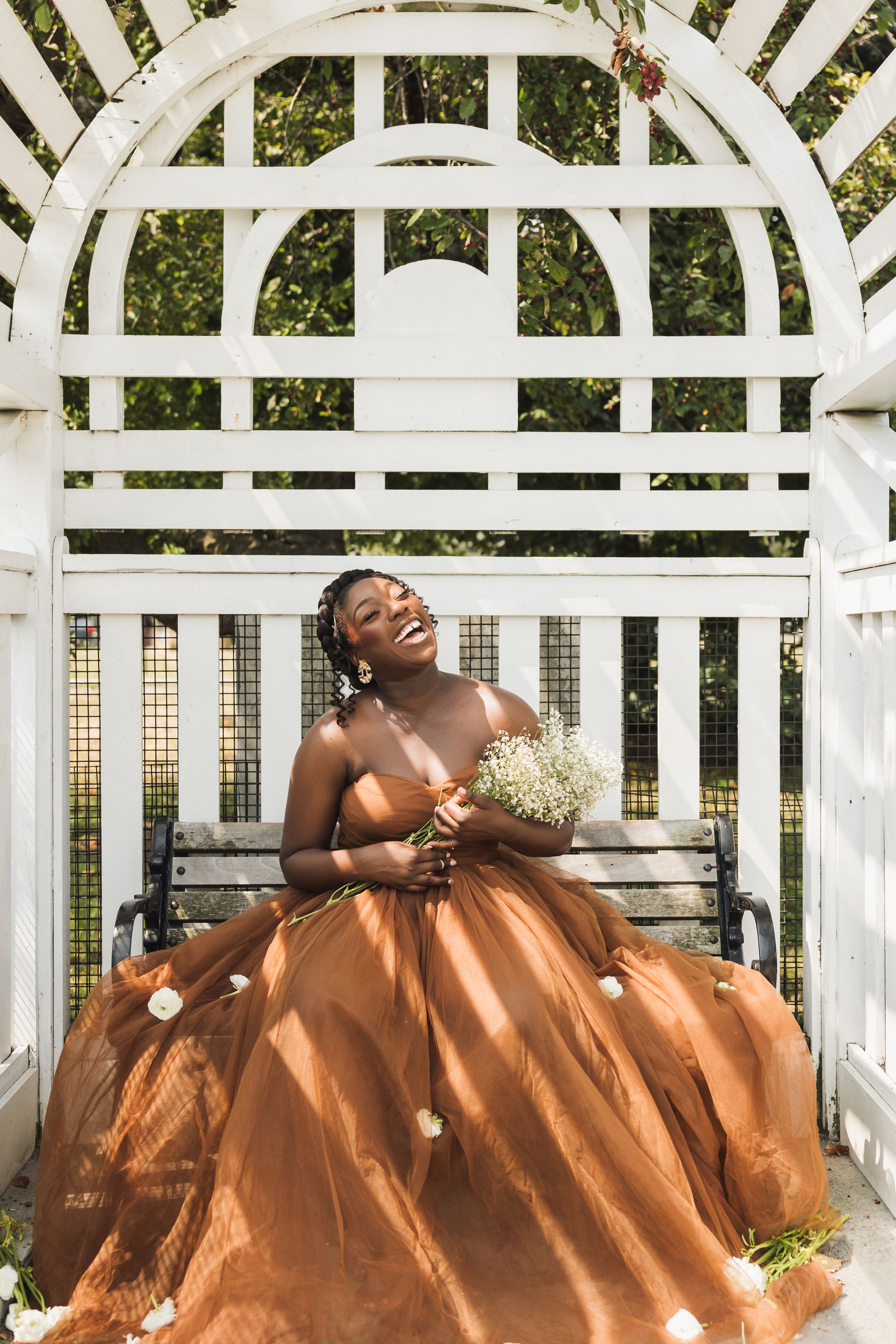 Person in flowing orange gown sits in white garden gazebo holding delicate flowers.