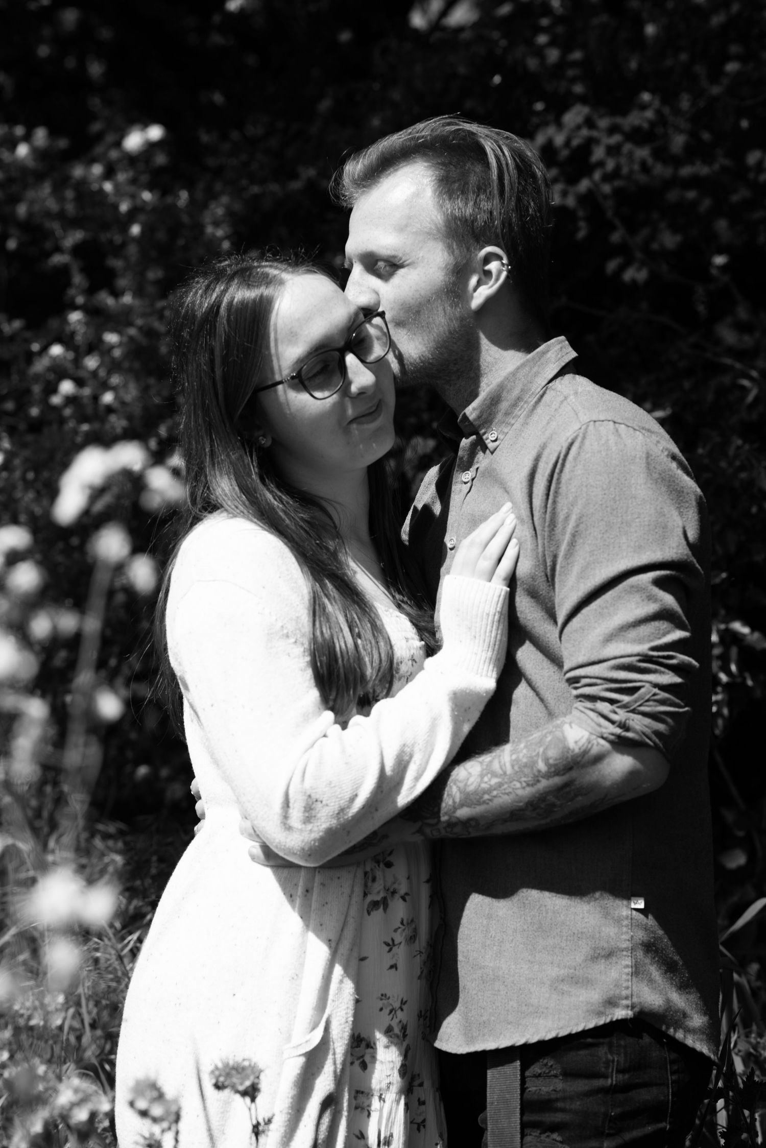 Couple in casual attire embracing while sitting on a tree branch in a green outdoor setting.