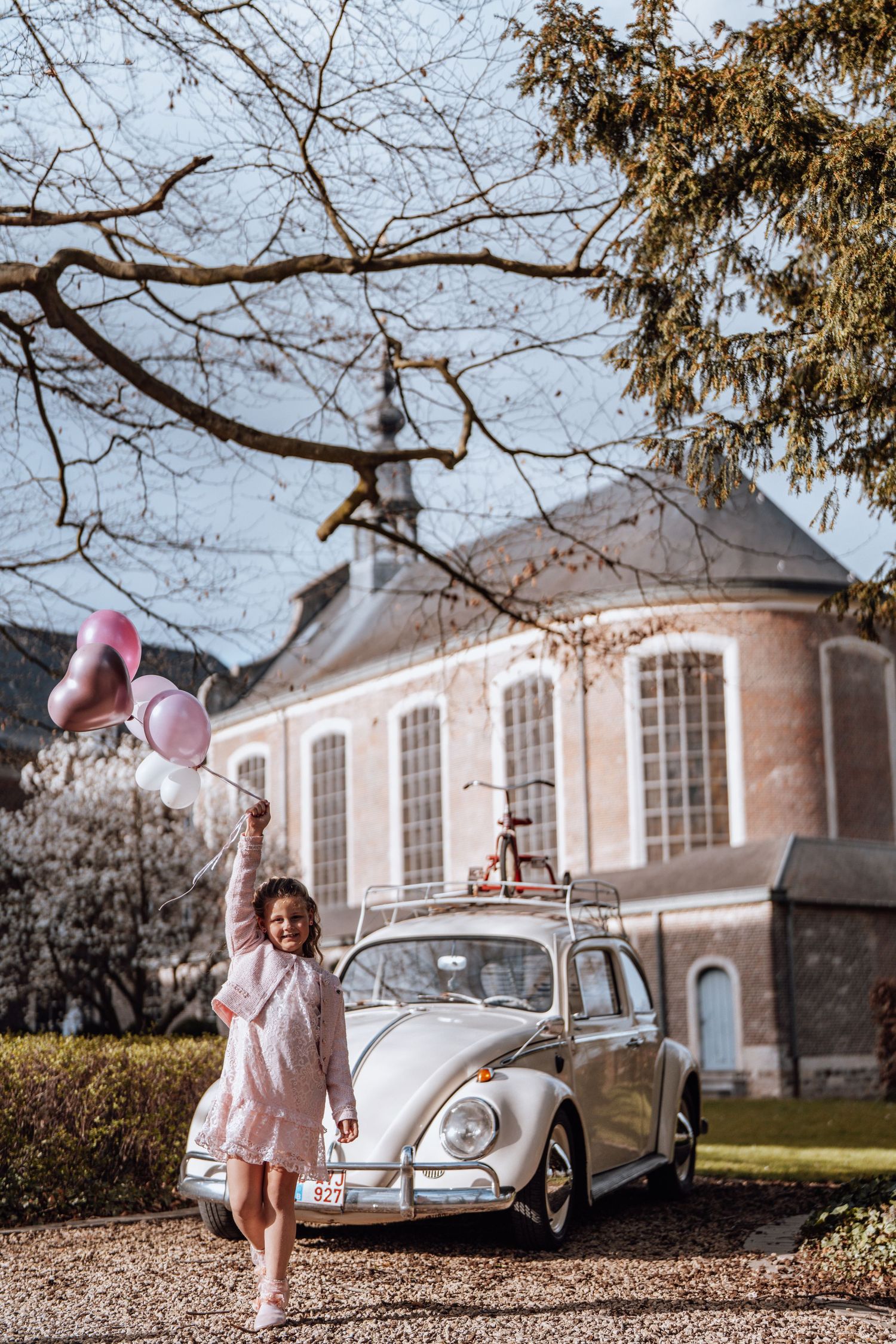 Een vintage Volkswagen kever voor een groot historisch gebouw met een meisje met roze hartvormige ballonnen in haar hand.