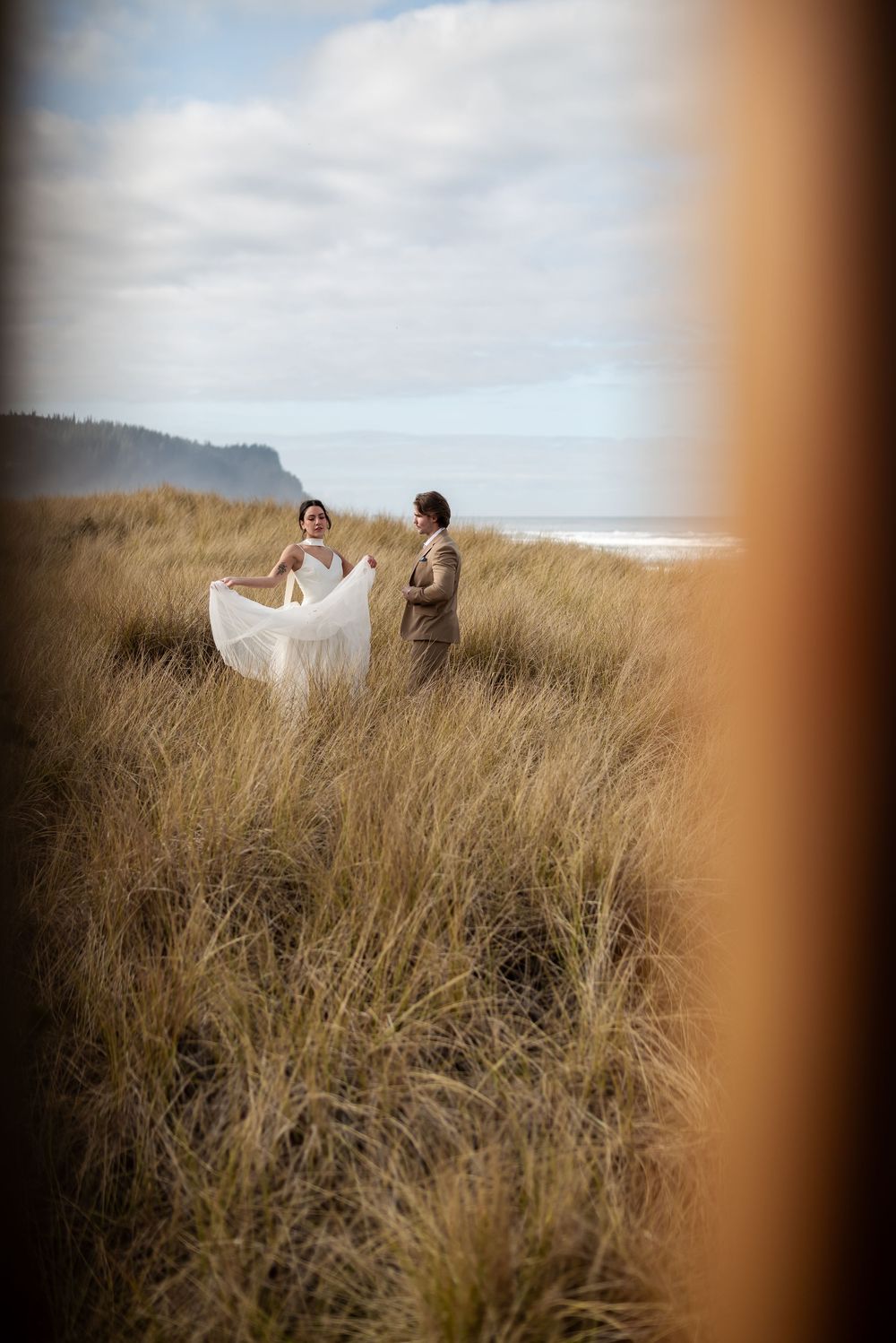 A couple in wedding attire standing in tall grass with ocean views.