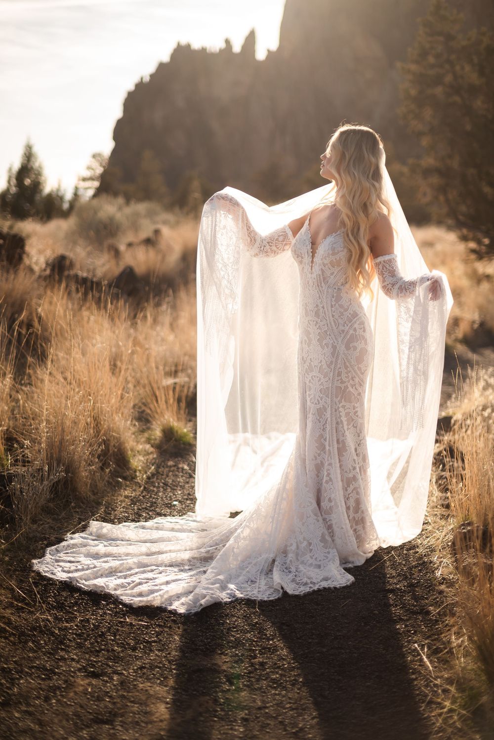 A flowing white lace wedding dress catches golden sunlight in a field of tall grass during sunset.
