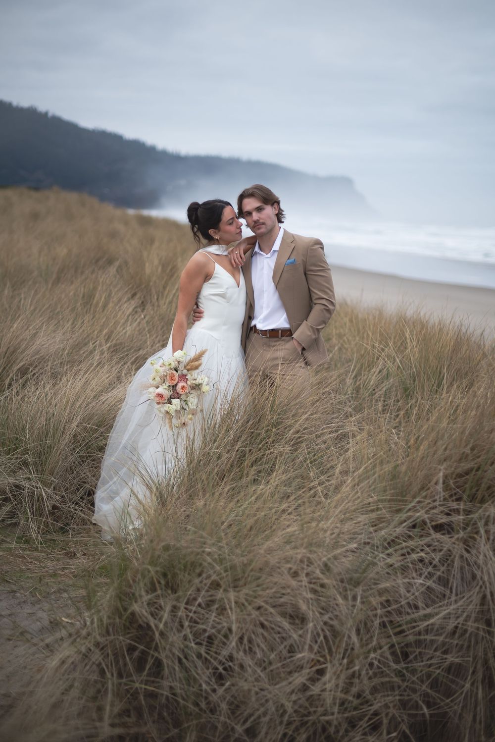 Wedding couple embracing in coastal dunes with overcast sky and ocean backdrop.