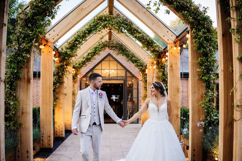A bride and groom share a tender moment under a wooden archway adorned with greenery and twinkling lights at Bartle Hall Wedding Venue