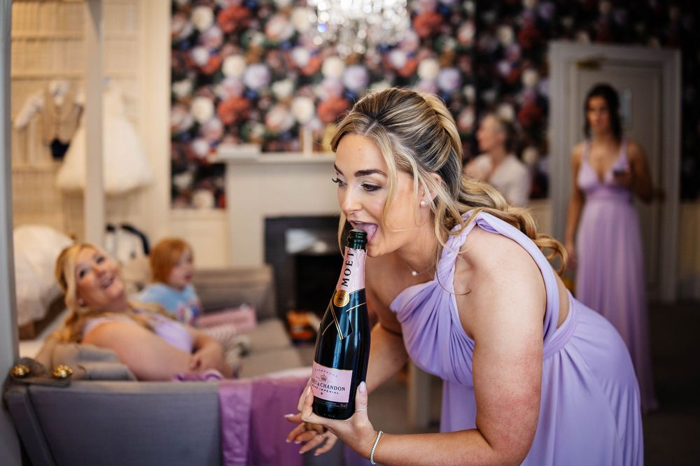 Bridesmaid in lavender dress captured at Bartle Hall, Lancashires 18th century house, share laughter as she drinks from Moet bottle