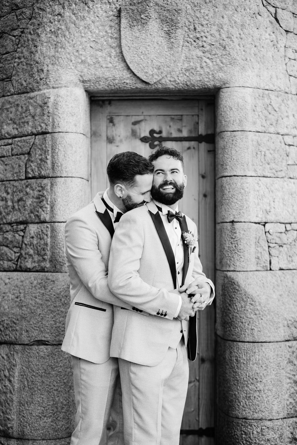 Two grooms embracing and smiling on their wedding day near a stone wall captured by Liverpool Wedding Photographer Lynette Matthews