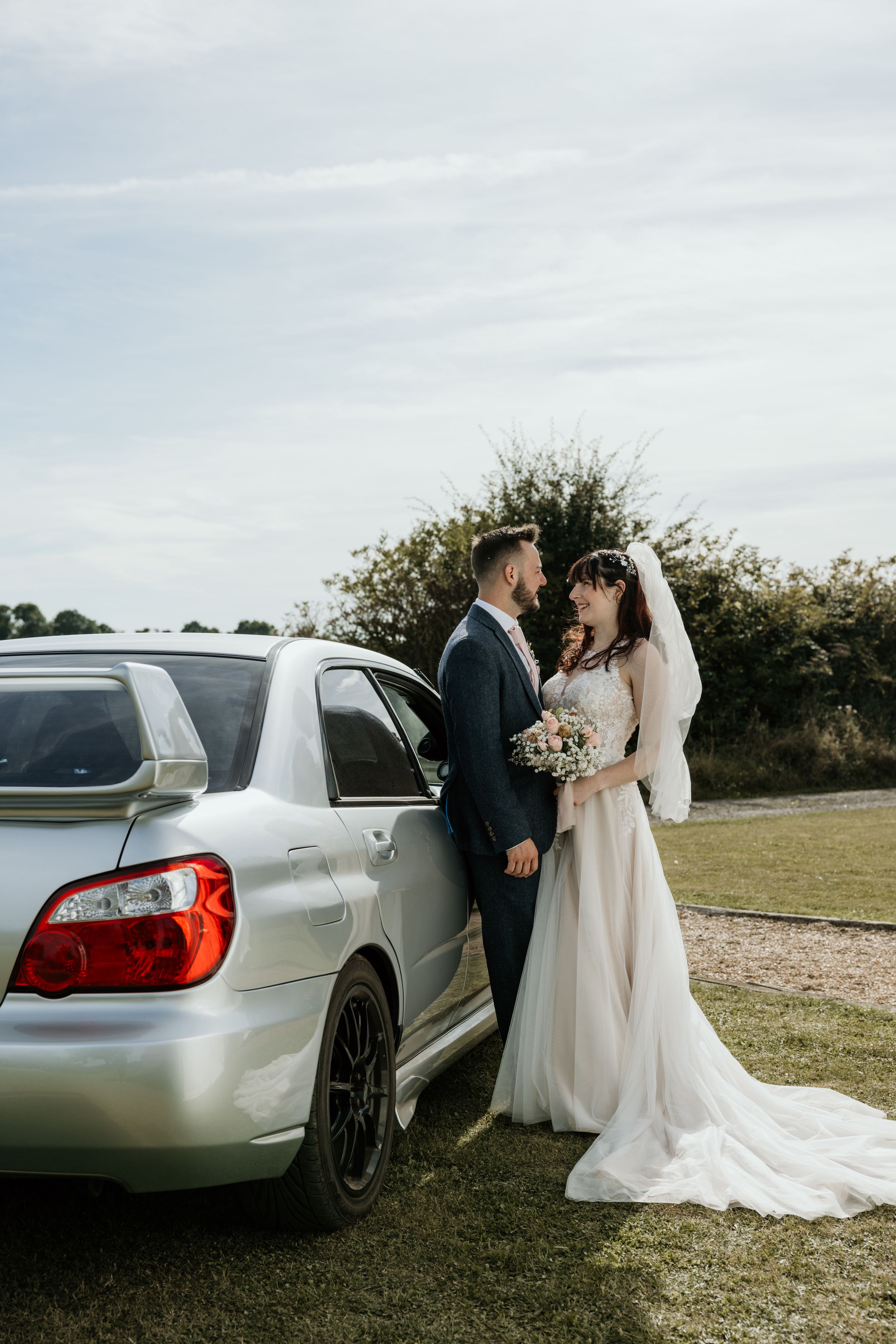A couple poses beside a silver car during their wedding photoshoot in a grassy field.