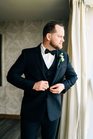 Groom in classic black tuxedo adjusting jacket by window.