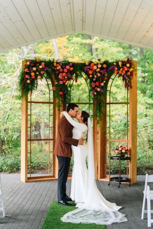 A couple embraces under a wooden arch decorated with vibrant orange and red flowers during an outdoor wedding ceremony.