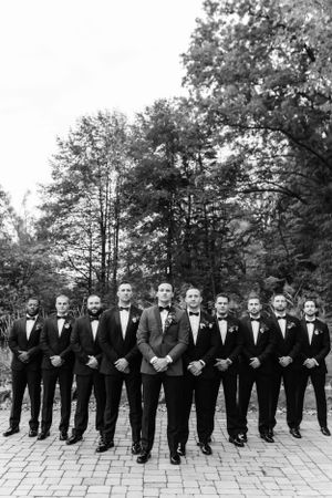 A formal wedding party of groomsmen in black tuxedos pose on a brick patio against a wooded backdrop.