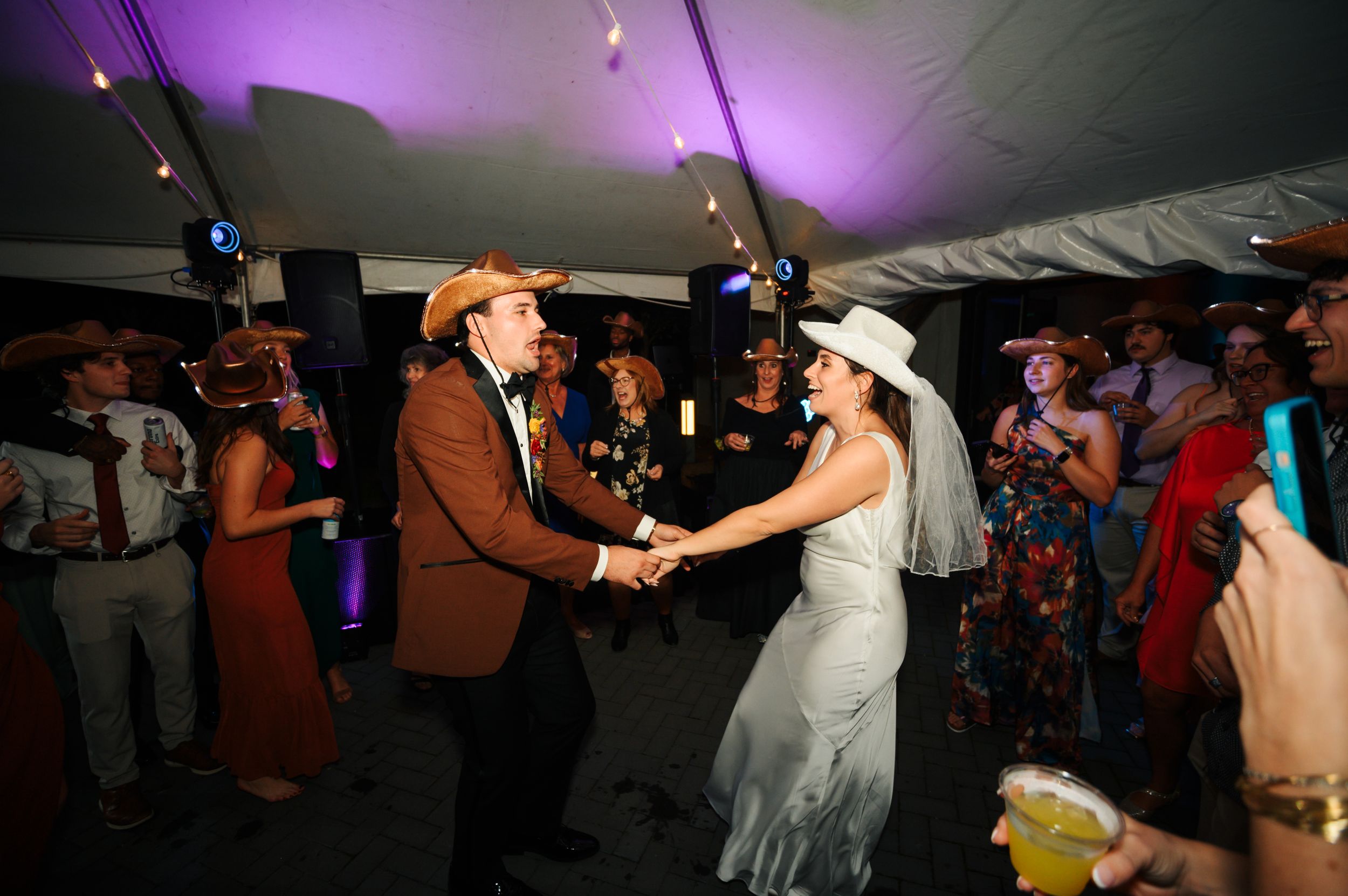 A lively dance scene at a wedding reception under a tent with purple lighting.