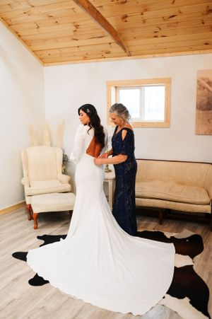 A bride in a white gown with long train being helped by someone in a navy dress in a rustic room.