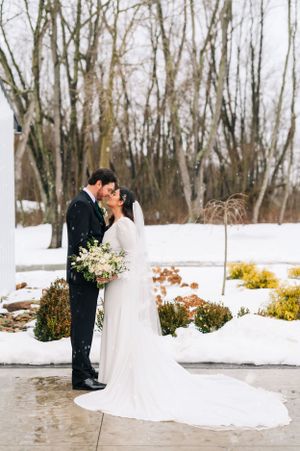 A winter wedding photo with a couple embracing on a snowy pathway surrounded by bare trees.