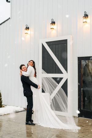 A joyful wedding couple embraces in front of a white barn with glowing lanterns during snowfall.