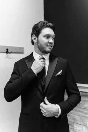 A formal portrait of a groom adjusting his tie while wearing a classic black tuxedo.