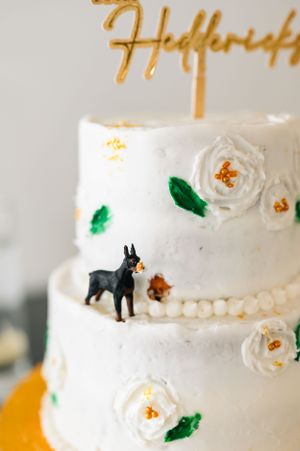 A white two-tier wedding cake decorated with sugar flowers and a small black dog figurine.