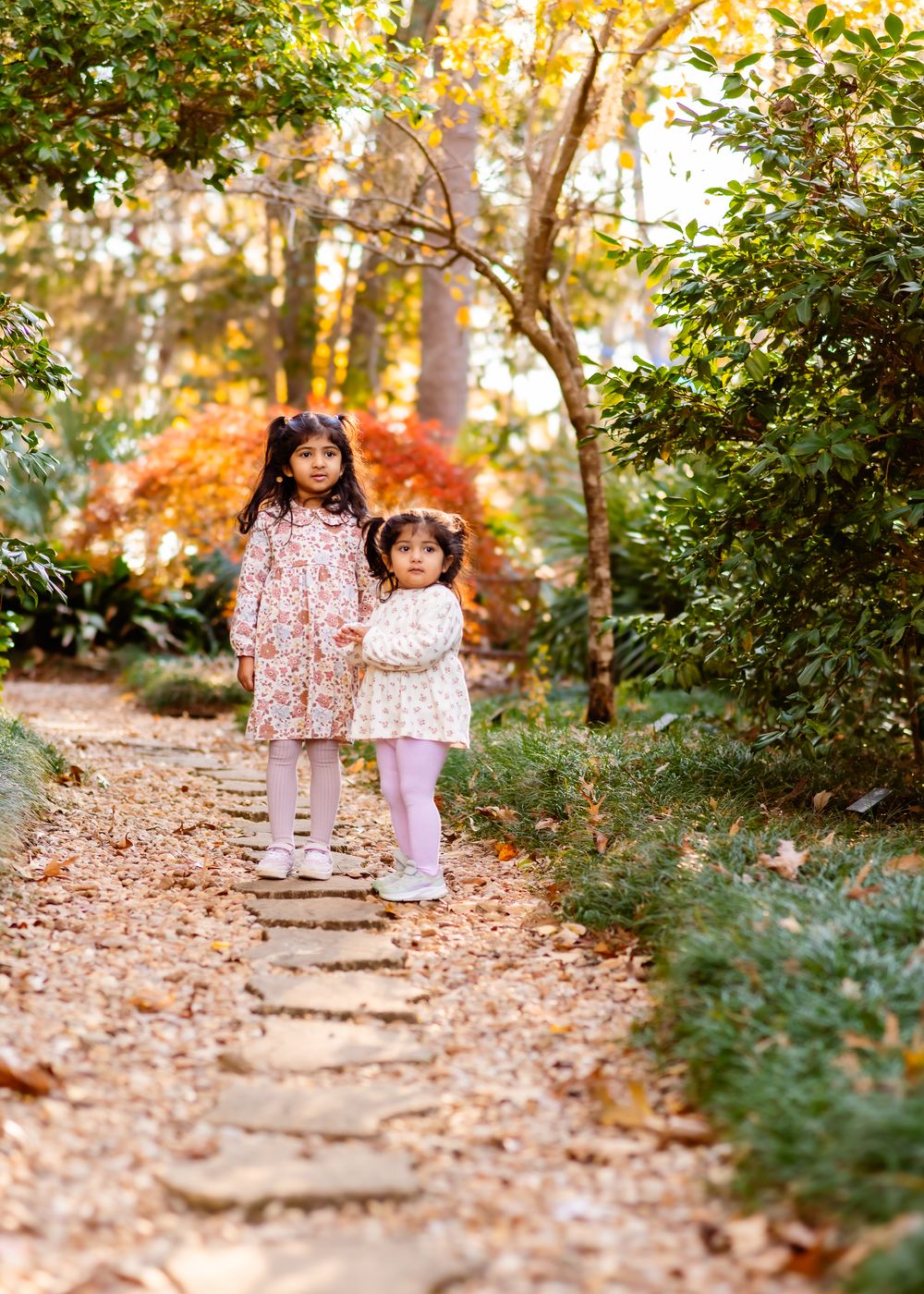 Two children walk down a leaf-covered woodland path during autumn while wearing matching floral dresses.