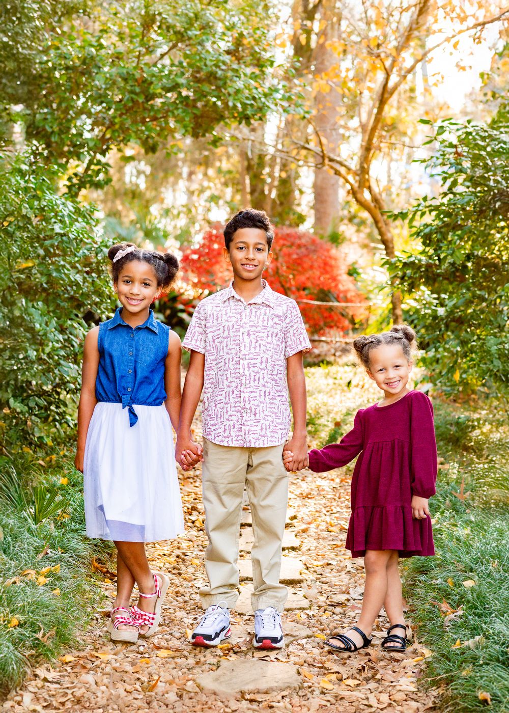 Three children hold hands while walking along a wooded path in summer.