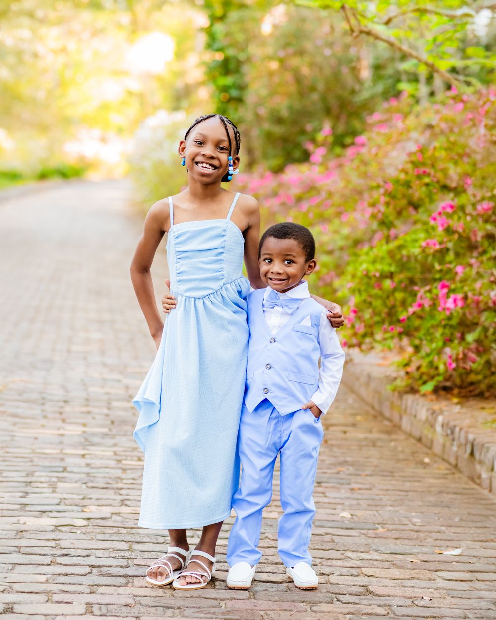 Two children in matching light blue outfits pose on a brick path surrounded by pink flowers.