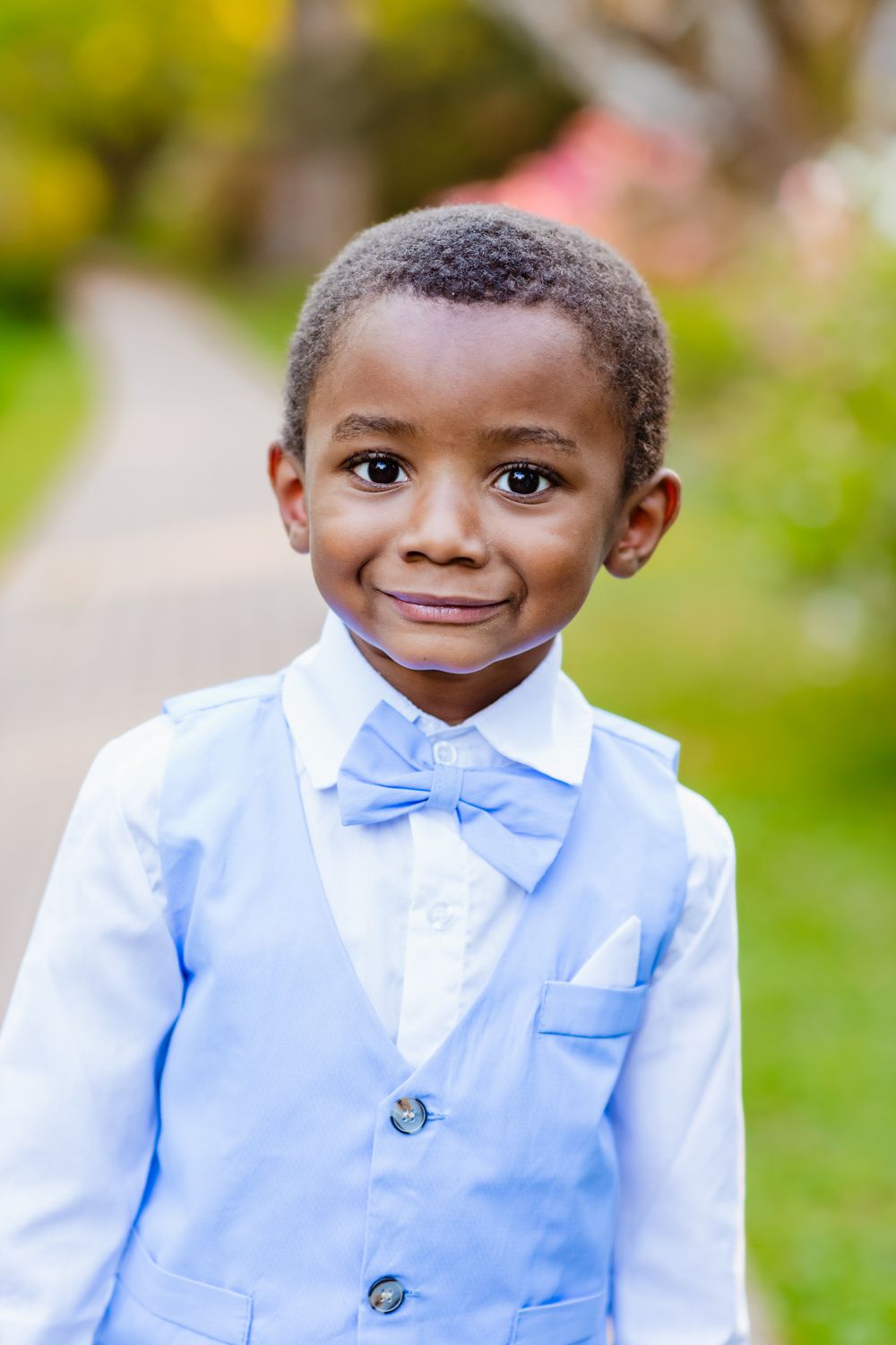 Adorable young boy in a light blue suit and bow tie smiling outdoors in a garden setting.
