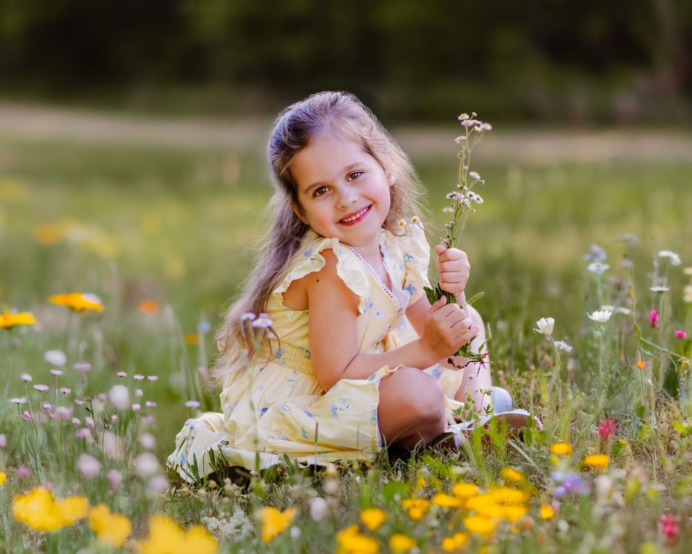 A smiling little girl in a yellow dress sits in a colorful wildflower meadow holding white flowers.