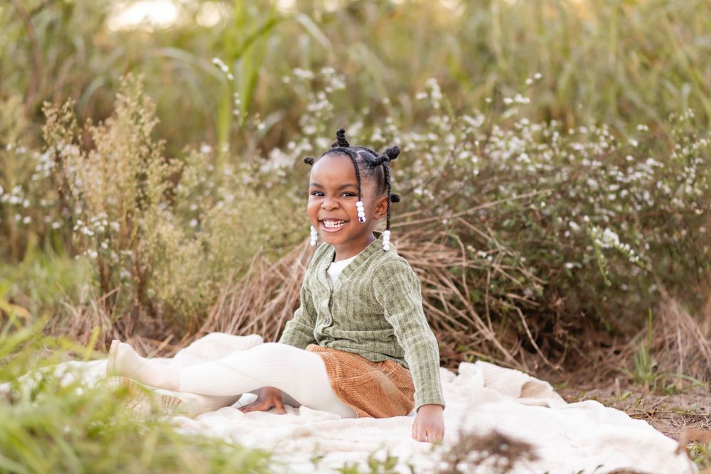 A small child wearing a knit sweater sits on a white blanket surrounded by flowering plants and tall grass.