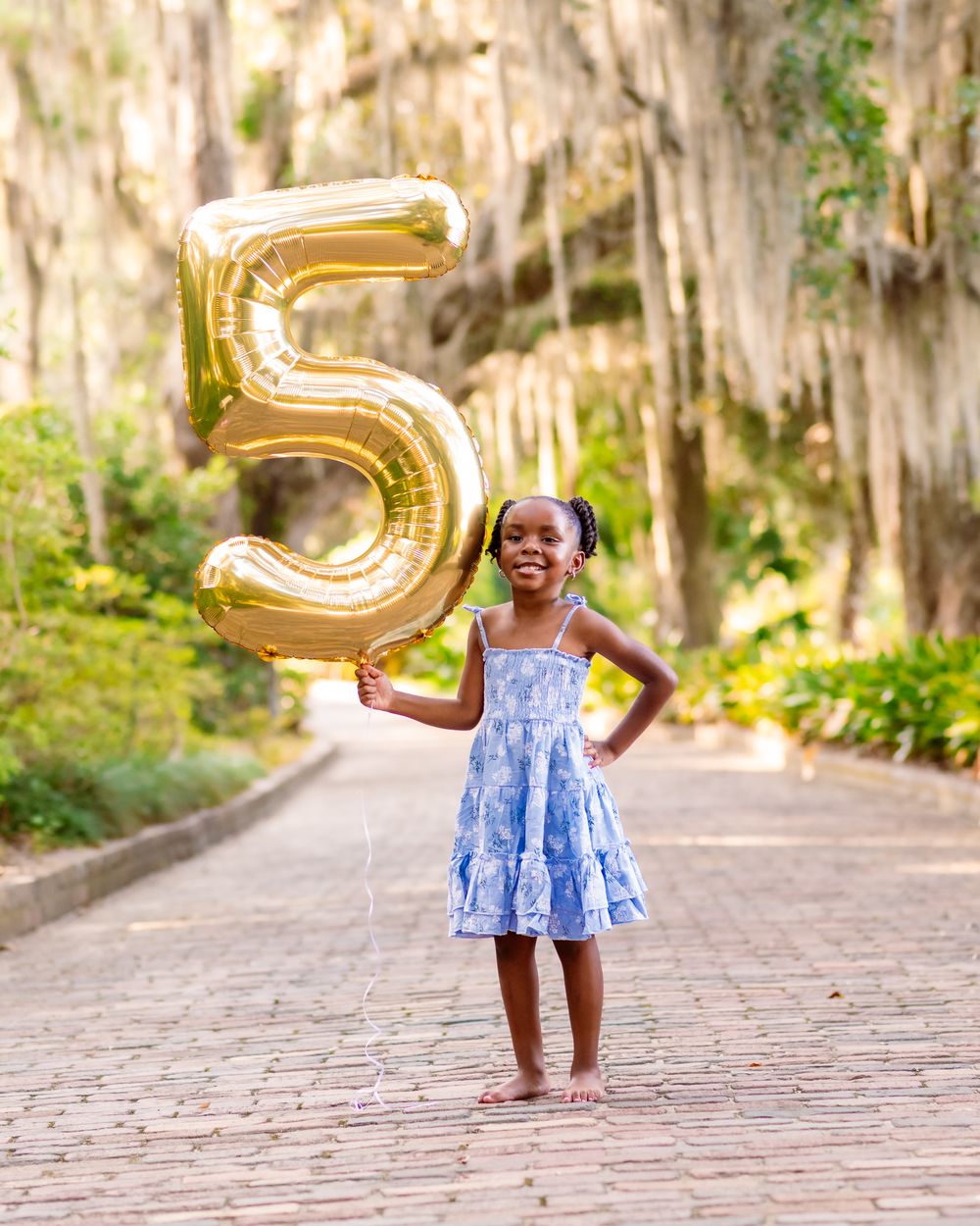 A young girl in a blue dress holds a gold number 5 balloon on a brick path, celebrating her 5th birthday outdoors.